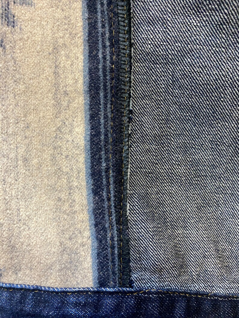 Op de afbeelding: Close-up van een denim naad met een contrasterend paneel van stof. De naad is gestikt met gele draad en de stof is een donkerblauwe denim.