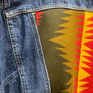 K&ouml;nnte beinhalten: Eine blaue Jeansjacke mit einem bunten, gemusterten Stoffpanel auf der R&uuml;ckseite. Das Panel zeigt ein geometrisches Design in Rot-, Gelb-, Gr&uuml;n- und Braunt&ouml;nen.