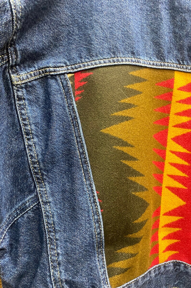 K&ouml;nnte beinhalten: Eine blaue Jeansjacke mit einem bunten, gemusterten Stoffpanel auf der R&uuml;ckseite. Das Panel zeigt ein geometrisches Design in Rot-, Gelb-, Gr&uuml;n- und Braunt&ouml;nen.