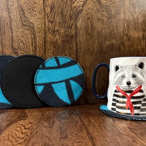 May include: A white ceramic mug with a raccoon design, black and white striped shirt, and a red scarf. The mug sits on a matching coaster. There are also three round coasters with blue and black patterns.