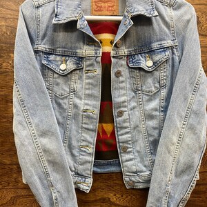 K&ouml;nnte beinhalten: Eine hellblaue Jeansjacke mit einem roten, gelben, braunen und schwarzen gemusterten Stofffutter. Die Jacke hat ein Levi Strauss & Co. Etikett mit der Gr&ouml;&szlig;e M.
