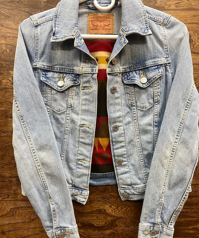 K&ouml;nnte beinhalten: Eine hellblaue Jeansjacke mit einem roten, gelben, braunen und schwarzen gemusterten Stofffutter. Die Jacke hat ein Levi Strauss & Co. Etikett mit der Gr&ouml;&szlig;e M.