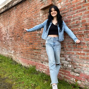 K&ouml;nnte beinhalten: Eine Frau tr&auml;gt eine hellblaue Jeansjacke, ein schwarzes Crop-Top, hellblaue Jeans mit Destroyed-Look und wei&szlig;e Sneaker. Sie steht vor einer Backsteinmauer.