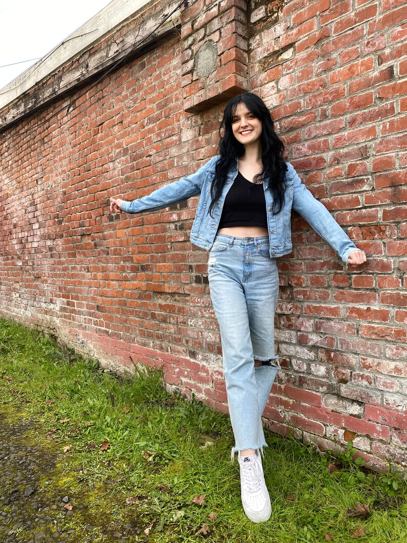 K&ouml;nnte beinhalten: Eine Frau tr&auml;gt eine hellblaue Jeansjacke, ein schwarzes Crop-Top, hellblaue Jeans mit Destroyed-Look und wei&szlig;e Sneaker. Sie steht vor einer Backsteinmauer.