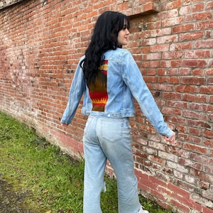 K&ouml;nnte beinhalten: Eine hellblaue Jeansjacke mit einem bunten Muster auf der R&uuml;ckseite. Das Muster zeigt Farbt&ouml;ne von Rot, Orange, Gelb, Braun und Schwarz. Die Jacke wird von einer Person getragen, die vor einer Backsteinmauer steht.