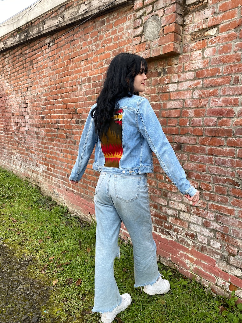 K&ouml;nnte beinhalten: Eine hellblaue Jeansjacke mit einem bunten Muster auf der R&uuml;ckseite. Das Muster zeigt Farbt&ouml;ne von Rot, Orange, Gelb, Braun und Schwarz. Die Jacke wird von einer Person getragen, die vor einer Backsteinmauer steht.