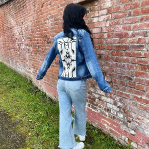 May include: A blue denim jacket with a white and black geometric pattern on the back. The jacket is worn by a person standing in front of a brick wall.