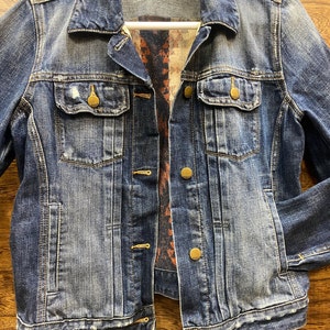 Op de afbeelding: Een blauwe denimjas met een geruite flanellen voering. De jas heeft een knoopsluiting en meerdere zakken. Het label op de jas staat "Gap 1969 m".