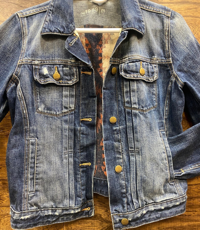Op de afbeelding: Een blauwe denimjas met een geruite flanellen voering. De jas heeft een knoopsluiting en meerdere zakken. Het label op de jas staat "Gap 1969 m".