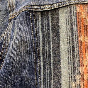 Op de afbeelding: Close-up van een denim jack met een gestreept en gepatroneerd paneel van stof. De denim is donkerblauw en het paneel is een mix van blauw, grijs en oranje.