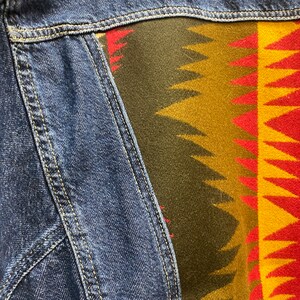 K&ouml;nnte beinhalten: Eine blaue Jeansjacke mit einem bunten, gemusterten Stoffpanel auf der R&uuml;ckseite. Das Panel zeigt geometrische Formen in Rot-, Gelb-, Gr&uuml;n- und Braunt&ouml;nen.