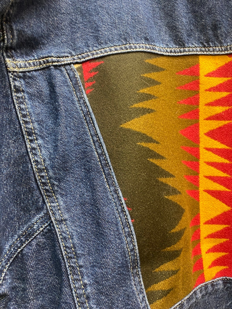 K&ouml;nnte beinhalten: Eine blaue Jeansjacke mit einem bunten, gemusterten Stoffpanel auf der R&uuml;ckseite. Das Panel zeigt geometrische Formen in Rot-, Gelb-, Gr&uuml;n- und Braunt&ouml;nen.
