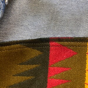 K&ouml;nnte beinhalten: Nahaufnahme einer Jeansjacke mit einem braunen, roten und gelben gemusterten Futter. Das Futter hat ein geometrisches Muster mit Dreiecken und einem dezenten Schimmer.