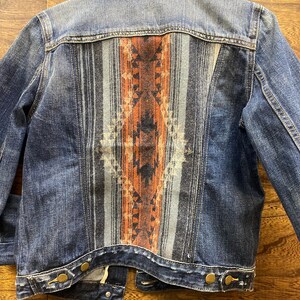 Op de afbeelding: Een donkerblauw denim jack met een gepatroneerd stoffen paneel op de rug. Het paneel heeft een geometrisch ontwerp in tinten oranje, blauw en zwart.