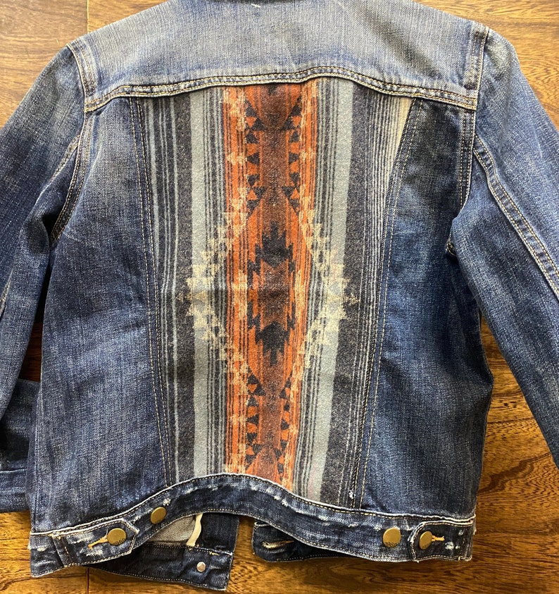 Op de afbeelding: Een donkerblauw denim jack met een gepatroneerd stoffen paneel op de rug. Het paneel heeft een geometrisch ontwerp in tinten oranje, blauw en zwart.