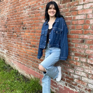 K&ouml;nnte beinhalten: Eine Frau tr&auml;gt eine blaue Jeansjacke, hellblaue Jeans mit einem zerrissenen Knie und wei&szlig;e Turnschuhe. Sie steht vor einer Backsteinmauer.