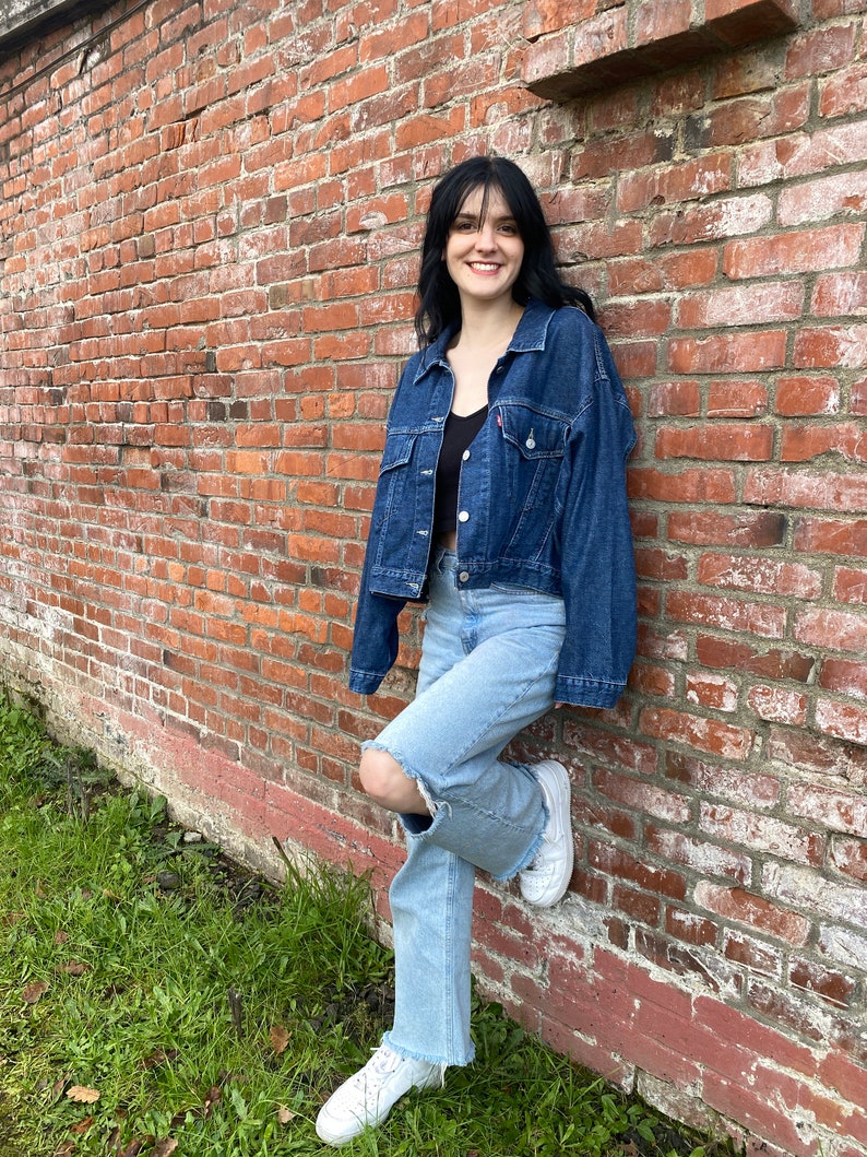 K&ouml;nnte beinhalten: Eine Frau tr&auml;gt eine blaue Jeansjacke, hellblaue Jeans mit einem zerrissenen Knie und wei&szlig;e Turnschuhe. Sie steht vor einer Backsteinmauer.
