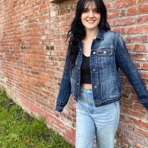 Op de afbeelding: Een vrouw draagt een donkerblauw denim jack over een zwart tanktop en lichtblauwe jeans met een gescheurde knie. Ze staat voor een bakstenen muur.