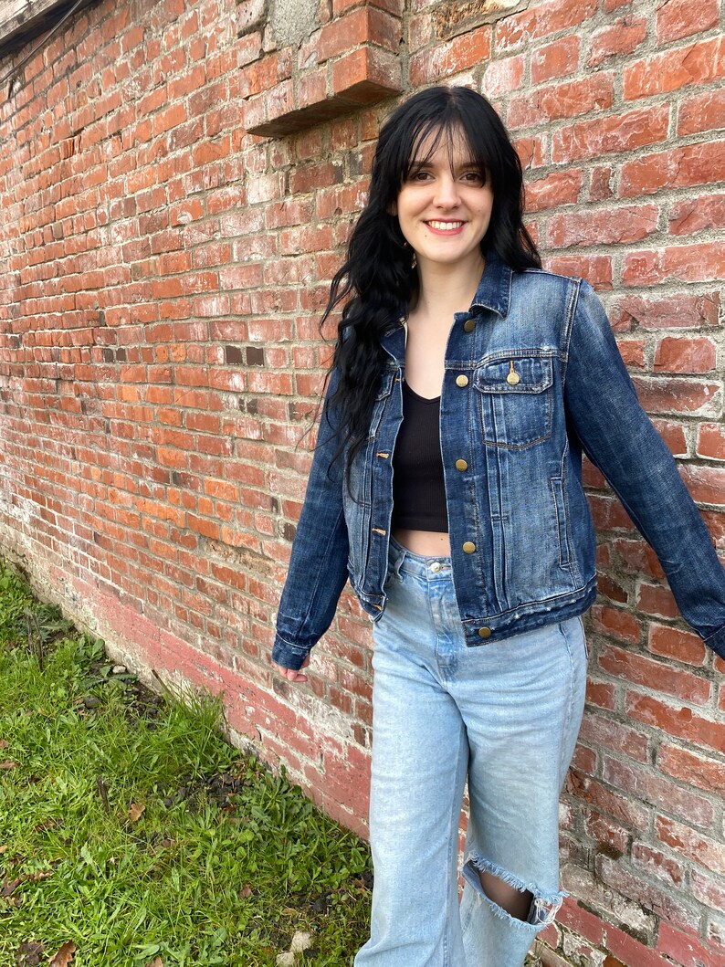 Op de afbeelding: Een vrouw draagt een donkerblauw denim jack over een zwart tanktop en lichtblauwe jeans met een gescheurde knie. Ze staat voor een bakstenen muur.