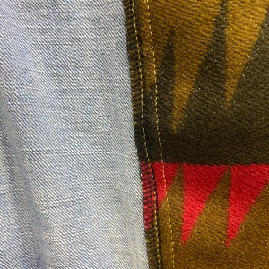 K&ouml;nnte beinhalten: Nahaufnahme einer Naht, an der zwei Stoffe zusammengen&auml;ht sind. Ein Stoff ist ein hellblauer Jeansstoff und der andere ein brauner, schwarzer, roter und gelber gemusterter Stoff.