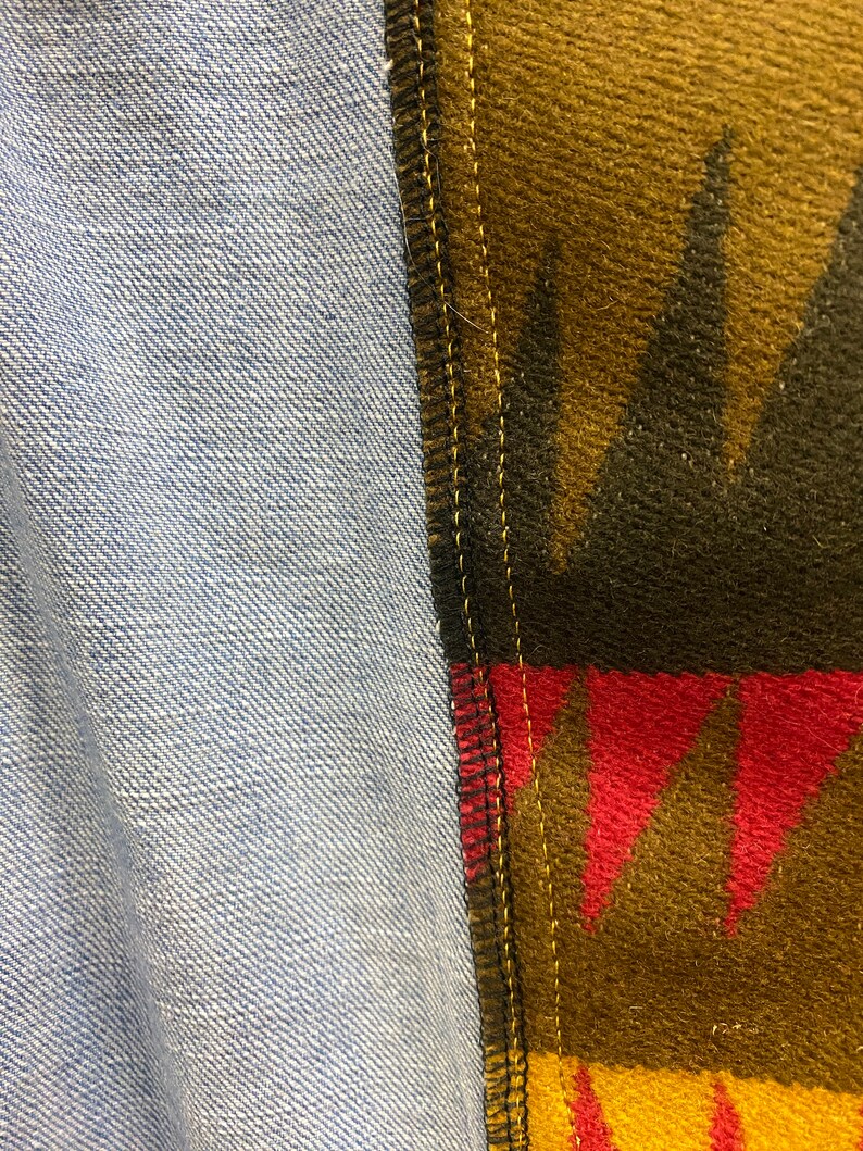 K&ouml;nnte beinhalten: Nahaufnahme einer Naht, an der zwei Stoffe zusammengen&auml;ht sind. Ein Stoff ist ein hellblauer Jeansstoff und der andere ein brauner, schwarzer, roter und gelber gemusterter Stoff.