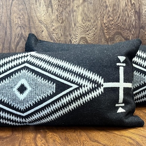 May include: Three dark gray rectangular throw pillows with a geometric white design. The pattern features a central diamond shape with a smaller diamond inside, and a cross-like design on the sides. The pillows are on a wooden surface.