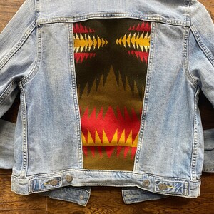 K&ouml;nnte beinhalten: Eine hellblaue Jeansjacke mit einem bunten geometrischen Muster auf der R&uuml;ckseite. Das Muster ist in Rot-, Gelb-, Gr&uuml;n- und Braunt&ouml;nen gehalten.