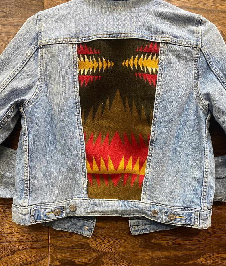 K&ouml;nnte beinhalten: Eine hellblaue Jeansjacke mit einem bunten geometrischen Muster auf der R&uuml;ckseite. Das Muster ist in Rot-, Gelb-, Gr&uuml;n- und Braunt&ouml;nen gehalten.