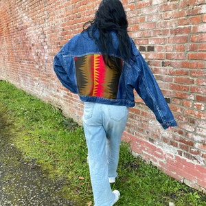 May include: A blue denim jacket with a colorful geometric pattern on the back. The jacket is worn by a person standing in front of a brick wall.