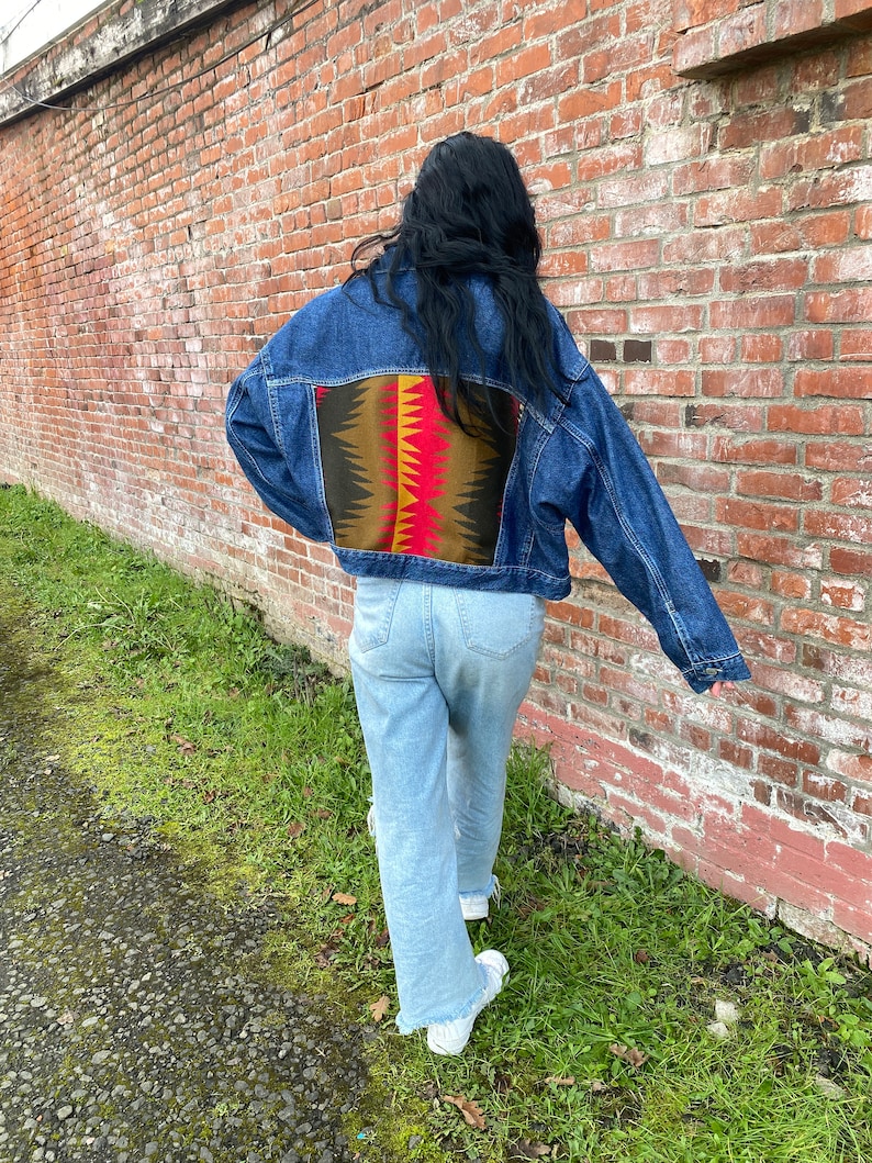 K&ouml;nnte beinhalten: Eine blaue Jeansjacke mit einem bunten geometrischen Muster auf dem R&uuml;cken. Die Jacke wird von einer Person getragen, die vor einer Backsteinmauer steht.