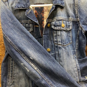 Op de afbeelding: Een blauwe denimjas met een bruine en blauwe gebloemde voering. De jas heeft een knoopsluiting, twee voorzakken en een enkele zak op de linkermouw. Het label op de jas luidt "Gap 1969 m".