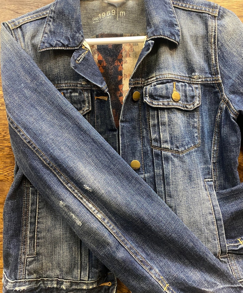 Op de afbeelding: Een blauwe denimjas met een bruine en blauwe gebloemde voering. De jas heeft een knoopsluiting, twee voorzakken en een enkele zak op de linkermouw. Het label op de jas luidt "Gap 1969 m".