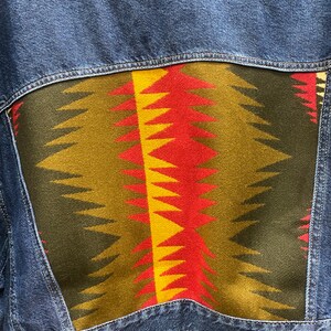 K&ouml;nnte beinhalten: Eine blaue Jeansjacke mit einem bunten geometrischen Muster auf der R&uuml;ckseite. Das Muster ist in Rot-, Gelb-, Gr&uuml;n- und Schwarzt&ouml;nen gehalten.