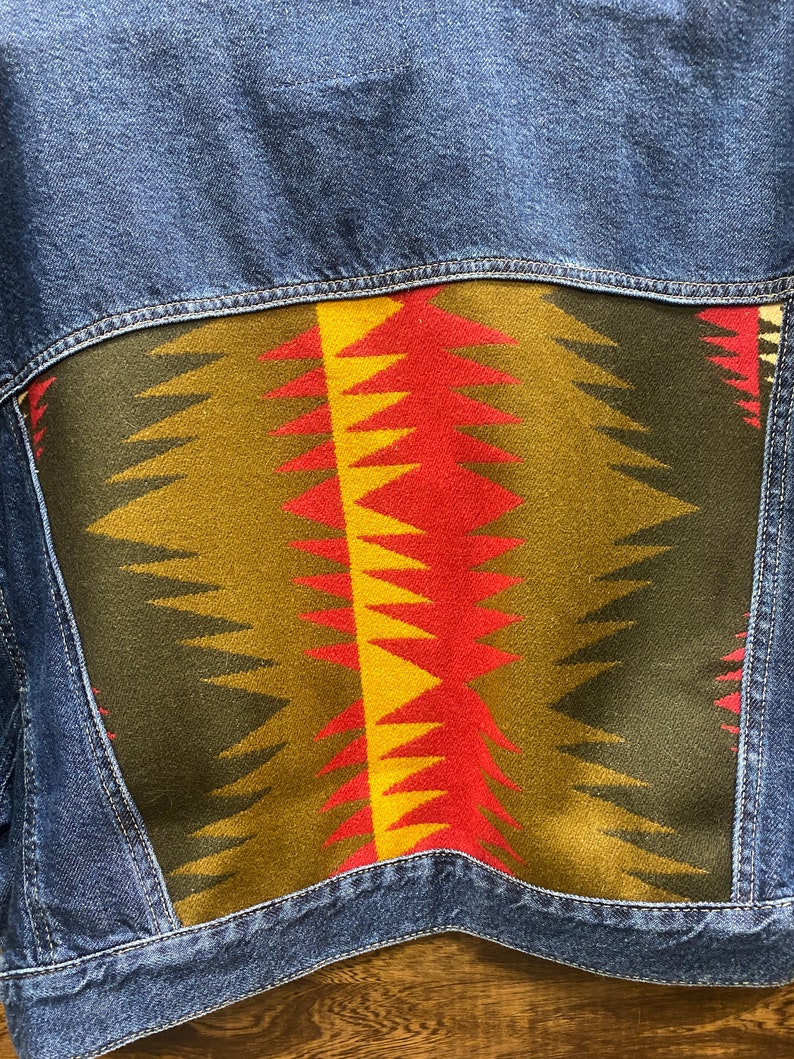K&ouml;nnte beinhalten: Eine blaue Jeansjacke mit einem bunten geometrischen Muster auf der R&uuml;ckseite. Das Muster ist in Rot-, Gelb-, Gr&uuml;n- und Schwarzt&ouml;nen gehalten.