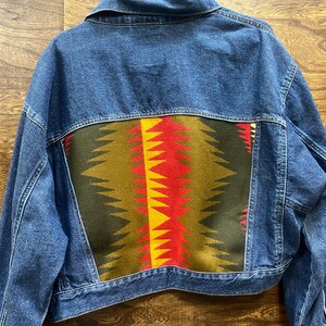 K&ouml;nnte beinhalten: Eine blaue Jeansjacke mit einem bunten geometrischen Muster auf der R&uuml;ckseite. Das Muster ist in Rot-, Gelb-, Gr&uuml;n- und Schwarzt&ouml;nen gehalten.