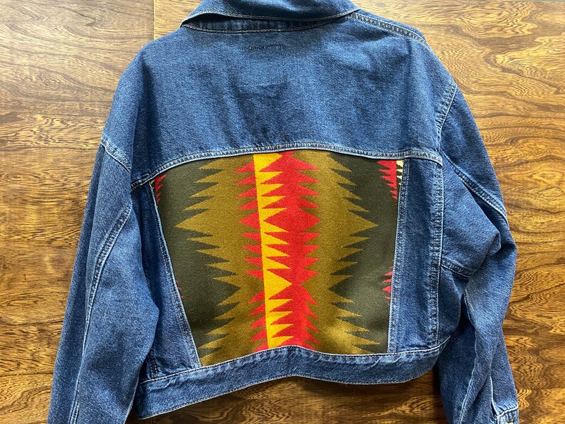 K&ouml;nnte beinhalten: Eine blaue Jeansjacke mit einem bunten geometrischen Muster auf der R&uuml;ckseite. Das Muster ist in Rot-, Gelb-, Gr&uuml;n- und Schwarzt&ouml;nen gehalten.