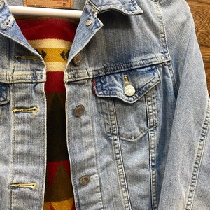 K&ouml;nnte beinhalten: Eine hellblaue Jeansjacke mit einem roten und gelben gemusterten Stofffutter. Die Jacke hat einen Knopfverschluss und eine kleine Tasche auf der Brust. Das Etikett auf der Jacke lautet "Levi Strauss & Co."