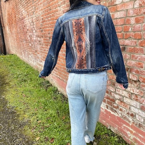 Op de afbeelding: Een blauwe denimjas met een kleurrijk patroon op de achterkant. De jas wordt gedragen over een lichtblauwe jeans.