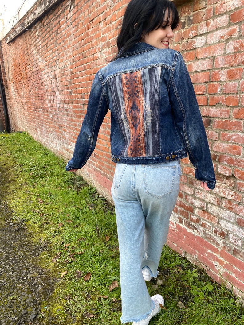 Op de afbeelding: Een blauwe denimjas met een kleurrijk patroon op de achterkant. De jas wordt gedragen over een lichtblauwe jeans.