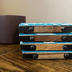 May include: A stack of five blue and white patterned coasters with wooden bases and black metal accents. Each coaster has a metal rivet on the side.