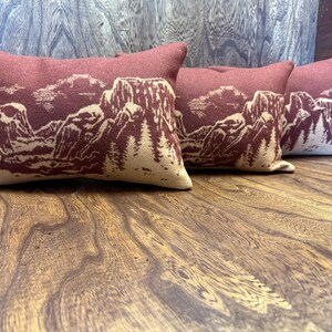 Puede incluir: Cuatro cojines rectangulares con un dise&ntilde;o de paisaje monta&ntilde;oso. Los cojines son de color rojo oscuro con un dise&ntilde;o de monta&ntilde;a y &aacute;rbol de color beige claro. Los cojines est&aacute;n colocados sobre una superficie de madera.