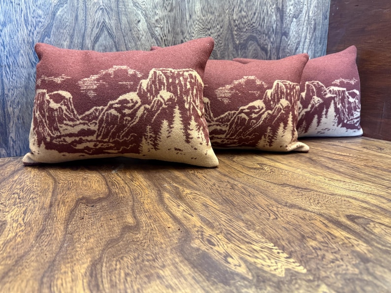 Puede incluir: Cuatro cojines rectangulares con un dise&ntilde;o de paisaje monta&ntilde;oso. Los cojines son de color rojo oscuro con un dise&ntilde;o de monta&ntilde;a y &aacute;rbol de color beige claro. Los cojines est&aacute;n colocados sobre una superficie de madera.