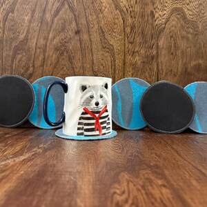 May include: A white ceramic mug with a raccoon design wearing a striped shirt and red scarf. The mug is surrounded by six round coasters, three with a black surface and three with a blue and gray pattern. The mug and coasters are on a wooden surface.