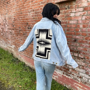 May include: Light blue denim jacket with a black and white geometric design on the back. The jacket has a classic collar, button-up front, and long sleeves. The person is wearing light blue jeans and white sneakers.