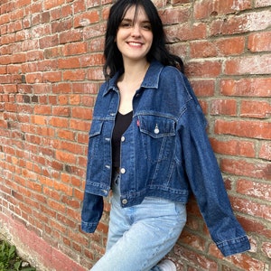 K&ouml;nnte beinhalten: Eine Frau tr&auml;gt eine dunkelblaue Jeansjacke &uuml;ber einem schwarzen Tanktop und hellblaue Jeans. Sie steht vor einer Backsteinwand.