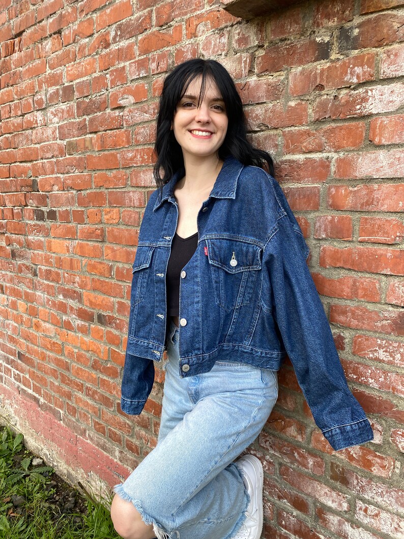 K&ouml;nnte beinhalten: Eine Frau tr&auml;gt eine dunkelblaue Jeansjacke &uuml;ber einem schwarzen Tanktop und hellblaue Jeans. Sie steht vor einer Backsteinwand.