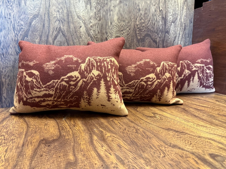 Puede incluir: Cuatro cojines rectangulares con un dise&ntilde;o de paisaje monta&ntilde;oso. Los cojines son de color rojo apagado con un dise&ntilde;o de monta&ntilde;a y &aacute;rboles beige. Los cojines est&aacute;n colocados sobre una superficie de madera.