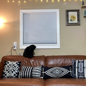 Puede incluir: Un sof&aacute; de cuero marr&oacute;n con cuatro cojines con dise&ntilde;os geom&eacute;tricos en blanco y negro. Un gato negro est&aacute; sentado en el sof&aacute;. Dos impresiones de girasoles enmarcadas y una estanter&iacute;a con adornos decoran la pared.