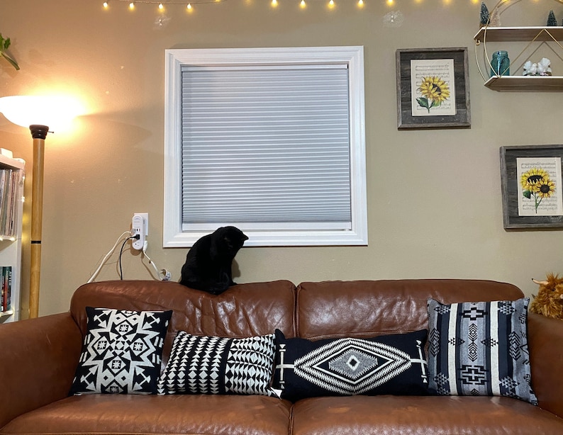 Puede incluir: Un sof&aacute; de cuero marr&oacute;n con cuatro cojines con dise&ntilde;os geom&eacute;tricos en blanco y negro. Un gato negro est&aacute; sentado en el sof&aacute;. Dos impresiones de girasoles enmarcadas y una estanter&iacute;a con adornos decoran la pared.