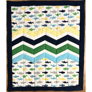 Puede incluir: Una colcha acolchada para bebé con borde azul marino. La colcha presenta un fondo blanco con coloridos estampados de tiburones. Un diseño de chevron en amarillo, azul marino, verde y azul claro añade un elemento decorativo.